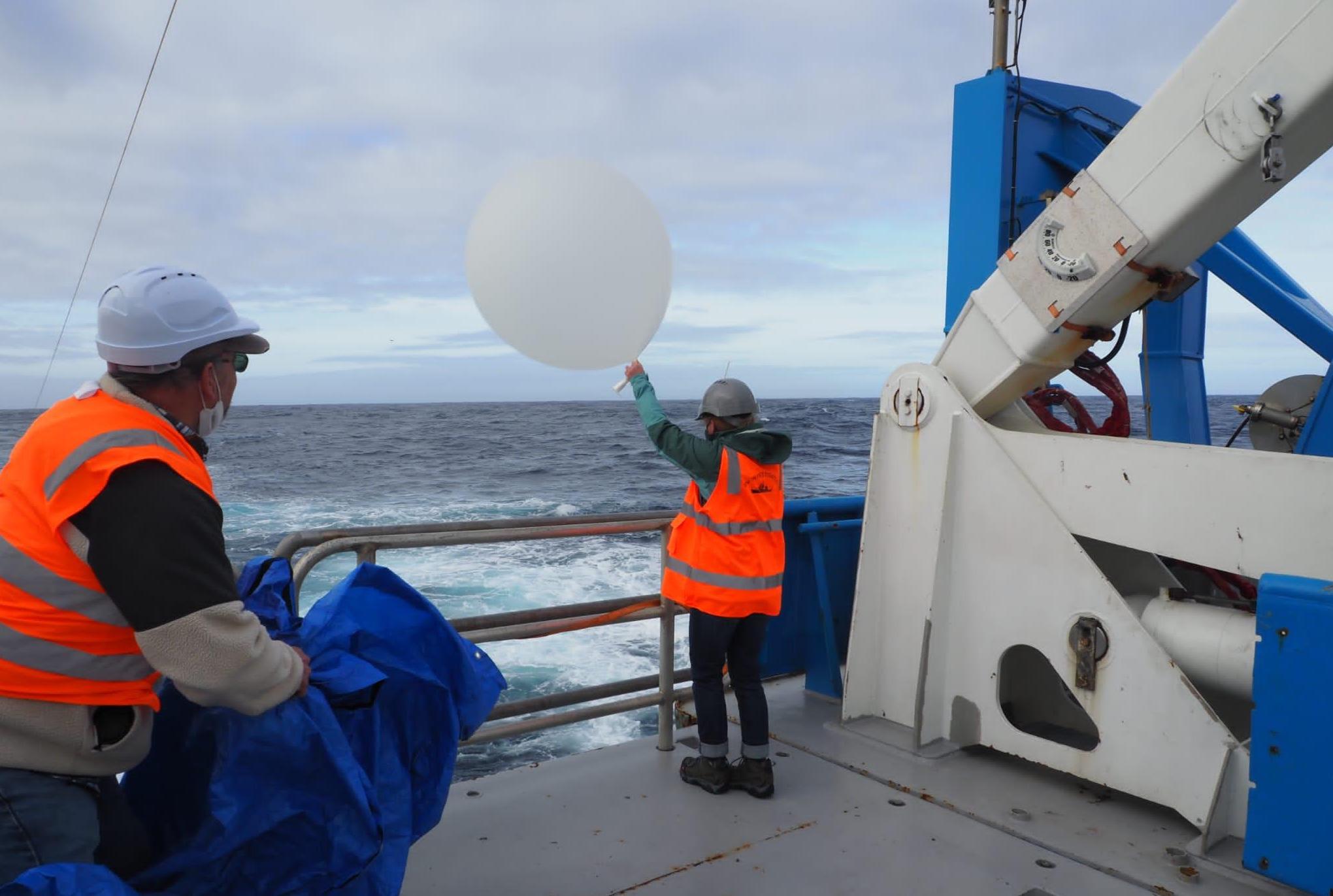 Kelsey stands at the ship's rail holding aloft a large white weather balloon, preparing to launch it into the atmosphere.